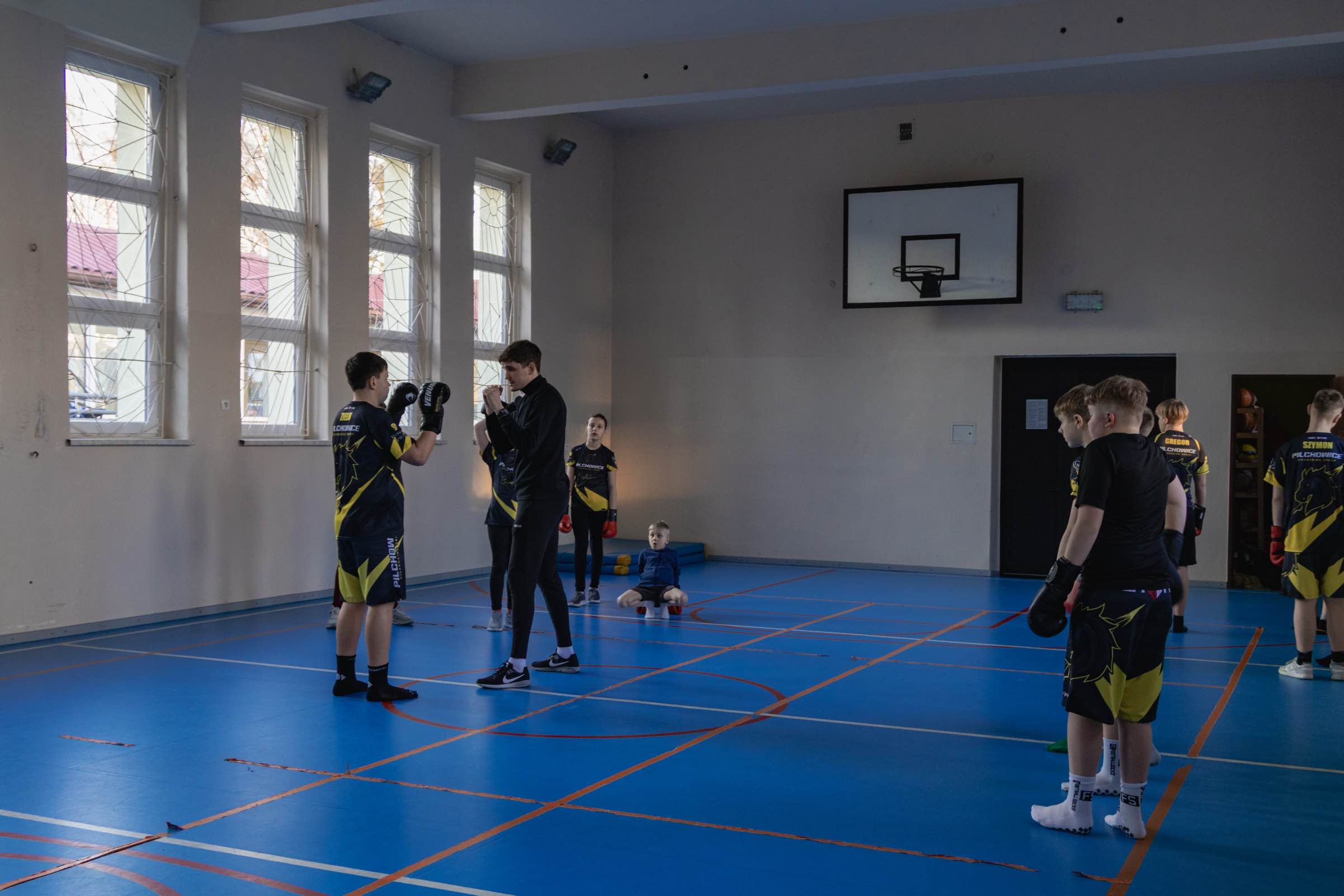 Szeroki kadr sali gimnastycznej, trener prowadzi rozgrzewkę z grupą